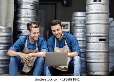 New order and production, sale of craft beer and small business. Smiling young attractive guys brewers in aprons sitting on floor in warehouse with metal barrels and looking at laptop, empty space - Powered by Shutterstock