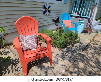 New Plastic Adirondack chairs located in a suburban backyard. One is blue and the other is orange. They have weatherproof pillows as a cushion, are next to a flower bed, and other outdoor essentials.