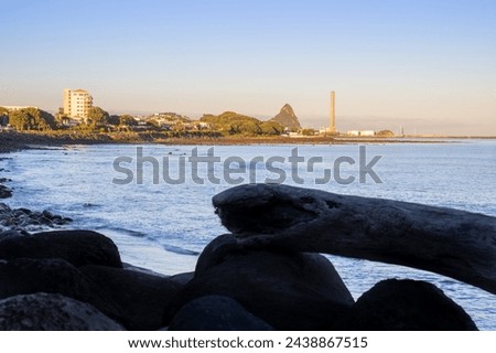 New Plymouth coastline view along Coastal Walkway beyond rocky foreshore and driftwood logs in foreground silhouette at sunrise looking down to port