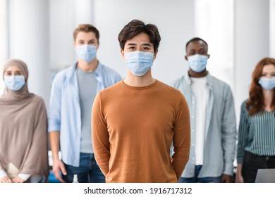 New Reality, Epidemic Concept. Portrait Of Asian Male Teen Wearing Protective Medical Mask Standing With Diverse Group Of International People In The Blurred Background, Posing And Looking At Camera