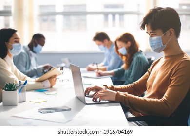 New Reality, People, Techology Concept. Group of diverse multicultural people wearing disposable surgical masks studying and working sitting at desk, writing and reading, asian guy using laptop