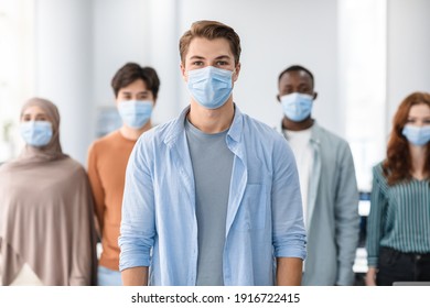 New Reality Rules, Epidemic Concept. Portrait Of Young Man Wearing Protective Medical Mask Standing With Diverse Group Of International People In The Blurred Background, Posing And Looking At Camera