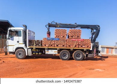 New Red Clay Bricks On Pallets Lifted By Crane Onto Truck Vehicle And Trailer For Delivery At Construction Building Site In Morning Blue Sky.