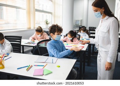 New Rules Due To Covid Pandemic. Female Teacher On School Nurse In Face Mask Helps Little Student With Sanitizing In The Classroom, Applying Alcohol Sanitizer Spray On Pupil's Hands. Personal Hygiene