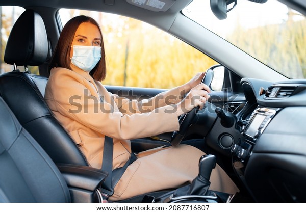 New social\
rules due to pandemic. Young woman wearing medical mask driving a\
car, female driver with covered face using protective measures for\
safety herself or\
passengers