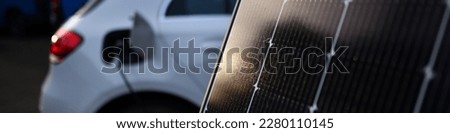 New solar panel with reflecting sun sharp in foreground and blurred used electric car plugged into charging socket in background. Standstill of the eco car in a wonderful evening atmosphere. 