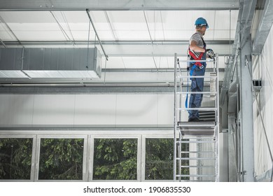 New Steel Warehouse Building And Interior Finishing By Caucasian Worker In His 40s On Aluminium Scaffolding. Commercial Building Interior.