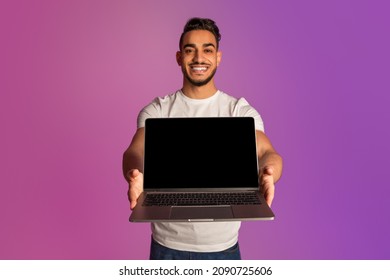 New Website. Happy Millennial Arab Man Holding Laptop Computer With Black Blank Screen, Showing Free Space For Your Mockup Design In Neon Light. Online Advertisement Concept