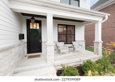 A new, white modern farmhouse with a dark wood door with windows, white pillars, a stone floor, and patio furniture.