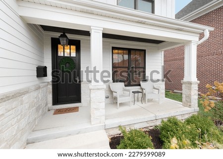 A new, white modern farmhouse with a dark wood door with windows, white pillars, a stone floor, and patio furniture.