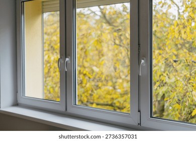 New window with a white plastic frame in a living space shows a view of trees with yellow leaves. Natural light enters the room, highlighting the comfortable space during fall. Empty windowsill.