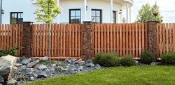 A New Wooden Fence With Stone Pillars Finished With Decorative Stones. Landscaping. Trimmed Lawn And Rock Hill. Young Maple Tree. Natural Materials. Solid Fence Wall. House Territory Accomplishment.