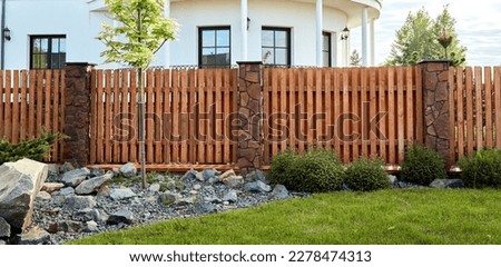 A new wooden fence with stone pillars finished with decorative stones. Landscaping. Trimmed lawn and rock hill. Young maple tree. Natural materials. Solid fence wall. House territory accomplishment.