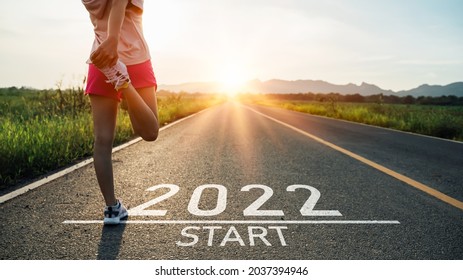 New Year 2022 Or Start Straight Concept.word 2022 Written On The Asphalt Road And Athlete Woman Runner Stretching Leg Preparing For New Year At Sunset.Concept Of Challenge Or Career Path And Change.