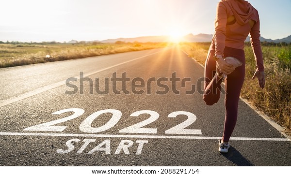 New year 2022 or start straight concept.word 2022\
written on the asphalt road and athlete woman runner stretching leg\
preparing for new year at sunset.Concept of challenge or career\
path and change.