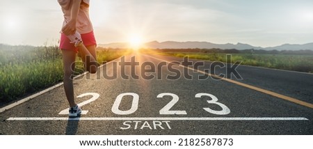 New year 2023 or start straight concept.word 2023 written on the asphalt road and athlete woman runner stretching leg preparing for new year at sunset.Concept of challenge or career path and change.