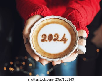 
New year 2024 celebrated coffee cup with number 2024 over frothy surface holding by female hands with nail polish and rings, dark background with blurred string lights. Holidays food art theme. - Powered by Shutterstock