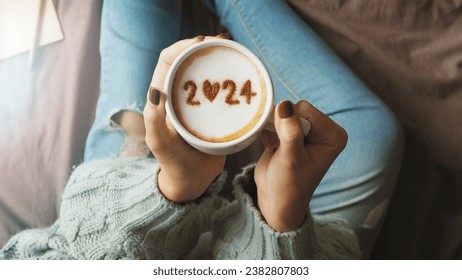 New year 2024 celebrated coffee cup with number 2024 on frothy surface of cappuccino in coffee cup holding by woman in green knitted sweater with jeans sitting on brown bed with notebook and pencil.