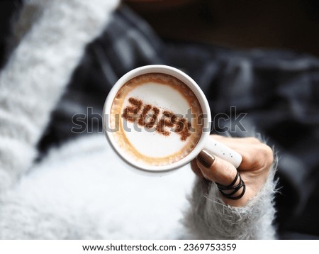 New year 2024 celebrated coffee cup with number 2024 over frothy surface holding by female hands with nail polish and ring on dark background. Holidays food art theme new year new you. (top view)