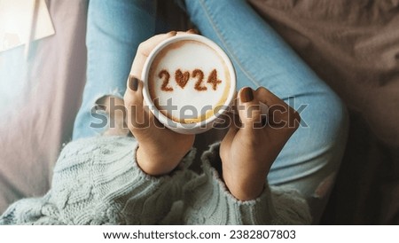 New year 2024 celebrated coffee cup with number 2024 on frothy surface of cappuccino in coffee cup holding by woman in green knitted sweater with jeans sitting on brown bed with notebook and pencil.