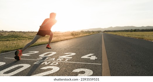 New year 2024 or start straight concept.word 2024 written on the asphalt road and athlete man runner stretching leg preparing for new year at sunset.Concept of challenge or career path and change.