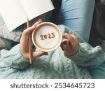 New year 2025 celebrated coffee cup with number 2025 on frothy surface of cappuccino in coffee cup holding by woman in green knitted sweater with jeans sitting on brown bed with pencil and notebook.