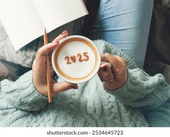 New year 2025 celebrated coffee cup with number 2025 on frothy surface of cappuccino in coffee cup holding by woman in green knitted sweater with jeans sitting on brown bed with pencil and notebook. - Powered by Shutterstock