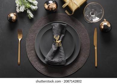New Year and Christmas table setting with black plates, gold deer ring, gift and gold cutlery. Golden gilded decorations on black background. View from above. Flat lay.