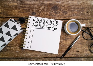 New Year Goals 2022 On Desk. 2022 Resolutions List With Notebook, Coffee Cup And Eyeglasses On Wooden Background. Goals, Plan, Strategy, Business, Idea, Action Concept