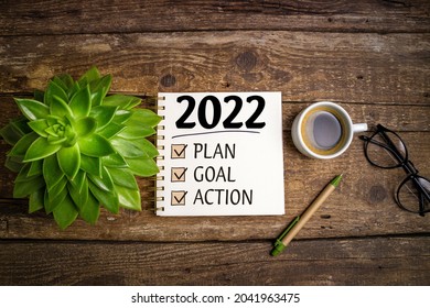 New year goals 2022 on desk. 2022 resolutions list with notebook, coffee cup and eyeglasses on wooden background. Goals, plan, strategy, business, idea, action concept. Top view