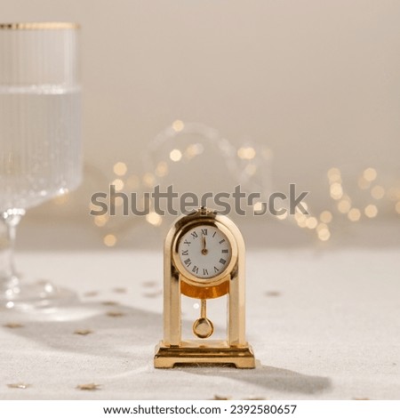 New Year holiday party celebration time. Vintage gold clock showing twelve o'clock, midnight, on beige linen tablecloth, blurred wineglass with sparkling wine, garland lights on background.
