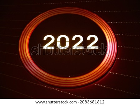 New Year neon sign on dark red background.  2022 glowing text in Christmas ball electric led lamp frame with light sparkles.  Celebration party, holiday greeting card design.