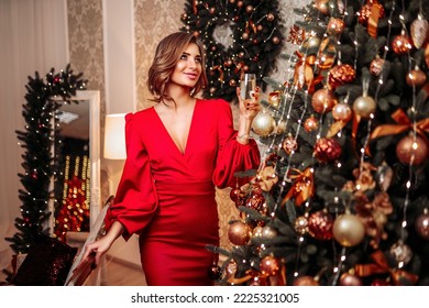 New Year Party, holidays, christmas and celebration concept. Beautiful happy girl in red dress with glass of sparkling wine near Christmas tree in decorated living room