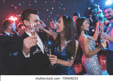 New Year Party. Young Couple Dancing With Glasses Of Champagne In Hands. Against The Background, Young People's Friends Are Dancing. Around Fly Confetti.