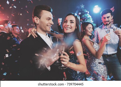 New Year Party. Young Couple Dancing With Glasses Of Champagne In Hands. Against The Background, Young People's Friends Are Dancing. Around Fly Confetti.