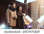 New Year Sales. Couple On Winter Shopping Holding Shopper Bags Looking At Clothing Store Window In Night City.