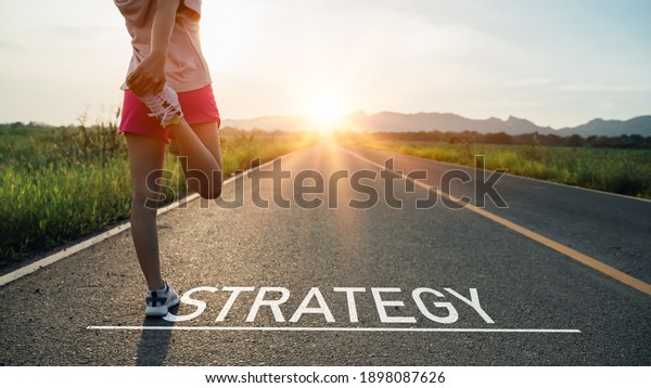 New year or start straight concept.word strategy\
written on the asphalt road and athlete woman runner stretching leg\
preparing for new year at sunset.Concept of challenge or career\
path and change