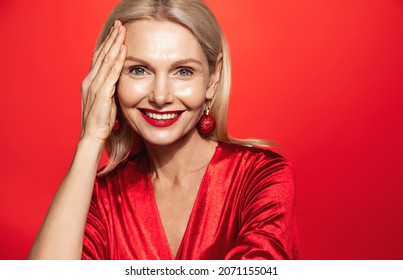 New Year Winter Holidays. Smiling Mature Woman With Red Lipsticks, Christmas Bubble Earrings And Elegant Dress, Touching Her Glowing Skin, Rejuvenation Concept