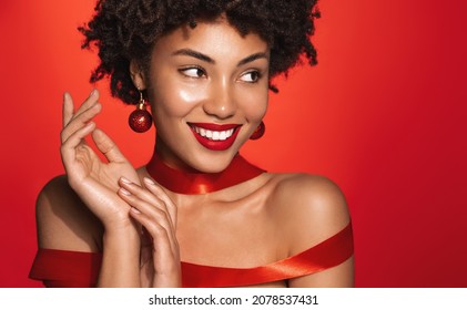 New Year And Women Beauty Concept. Beautiful Black Woman In Red Gift Ribbon, Christmas Tree Toy Earrings, Smiling And Looking Aside, Red Background