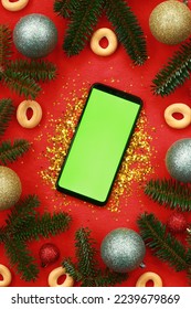 New Year's composition. A smartphone with a green screen, gold glitter, Christmas tree branches and decor on a red background. Christmas mockup for your advertisement