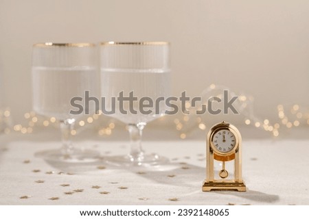 New Year's eve celebration concept, golden clock with twelve o'clock time, midnight, on beige table with gold star confetti, blurred wineglasses with sparkling wine and garland lights on background.