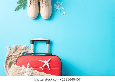 New Year's Flight Preparations: top view image of a red suitcase, miniature plane, winter essentials – boots, scarf, garnished with fir twigs and snowflakes against a vibrant blue backdrop