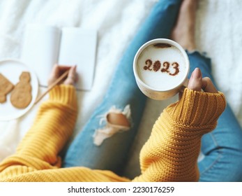New Year's goal setting, number 2023 on frothy surface of cappuccino in white coffee cup holding by woman in yellow knitted sweater with jeans sitting on bed while writing down her resolutions. - Shutterstock ID 2230176259