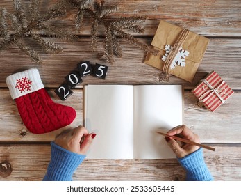 New Year's goal setting theme woman’s hand holding pencil to write down her resolutions on blank white notebook paper over wood plank table with black painted wooden dice with number 2025, stocking - Powered by Shutterstock