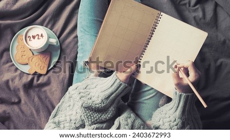 New Year's goal setting, woman in green knitted sweater with jeans write down her resolutions. Notebook and pencil in female hands holding over blurred coffee cup with number 2024 on frothy surface.