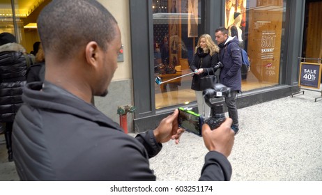 New York City - Circa 2017: Man walks down NYC Manhattan sidewalk using DJI Osmo 4K stabilizer camera recording on iphone app. Tourists use selfie stick in background to share photos on social media