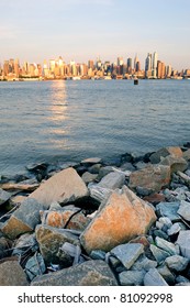 New York City Manhattan Midtown Skyline At Sunset With Reflection Over Skyscraper And River Viewed From New Jersey Hudson River Shore With Rocks.