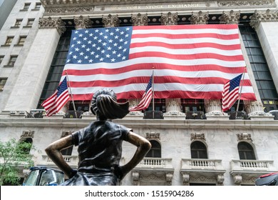 New York City/USA - September 2, 2019: Fearless Girl Standing Bronze Strong Looking Up At The Iconic Structure Of The NY Stock Exchange In Lower Manhattan With American Flag. 