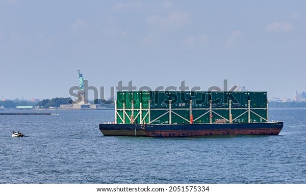 New York, NY - August 26,\
2021: A barge loaded with municipal-waste shipping containers\
moored in NY Harbor in front of The Statue of Liberty on a hot\
summer day. 