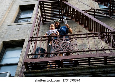 New York, NY / United States - May 16th 2020: A man and his toddler son ring a cowbell on their fire escape to thank medical workers in Washington Heights, Manhattan, New York City.